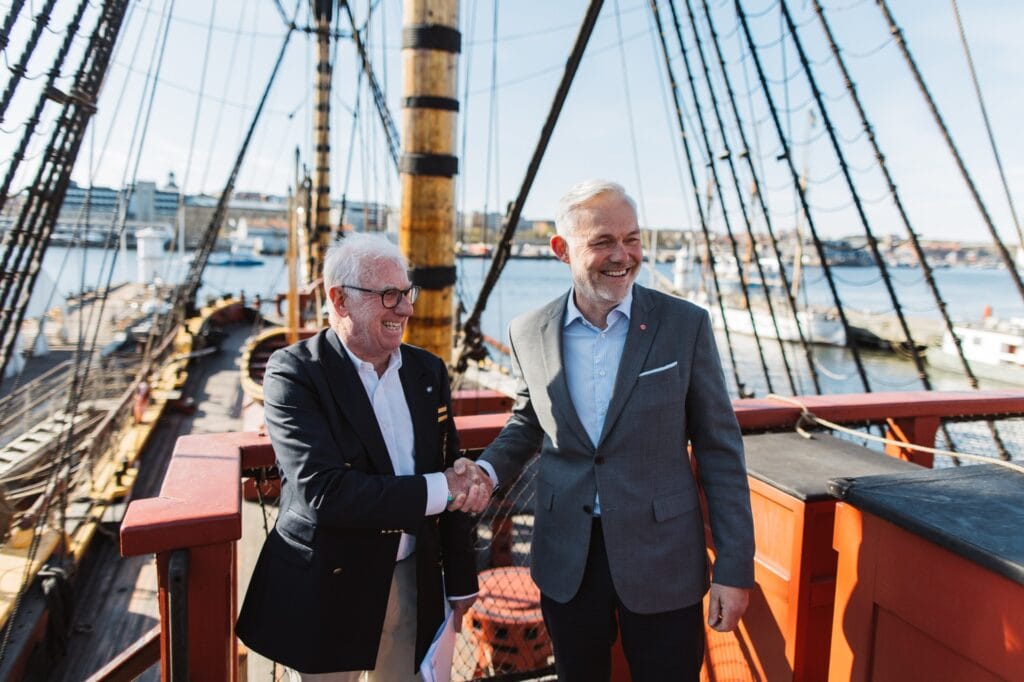 Two men in suits shake hands on a ship’s red railing with rigging and water in the background, smiling.