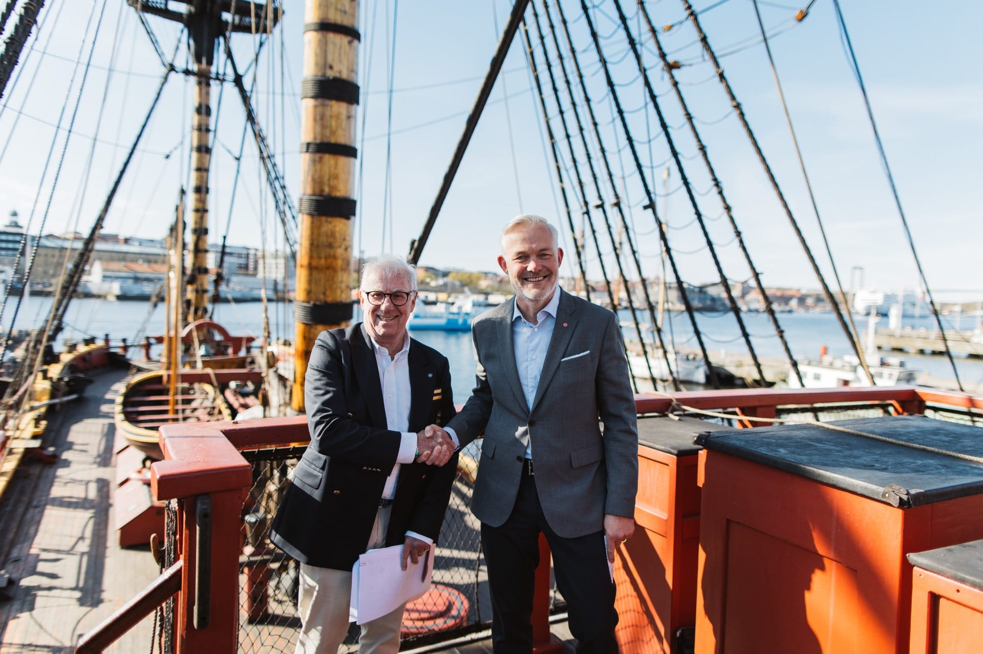 Two men in suits shake hands on a ship deck, papers in hand, with rigging and harbor in the background.