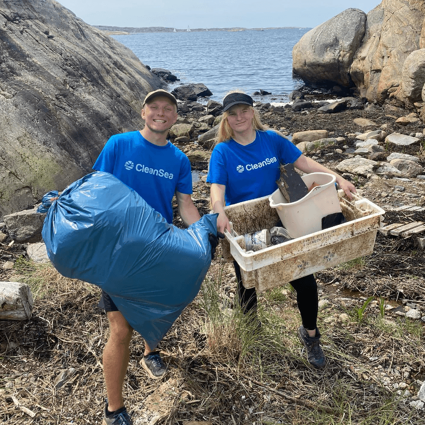 Two volunteers in blue CleanSea shirts collect litter on a rocky shoreline, holding a large blue trash bag and a tub of debris.