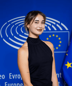 Julieta Martínez standing in front of a blue wall with the european flag behind. She is smiling to the camera.